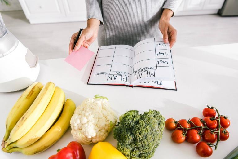 cropped view of woman near notebook with meal plan and vegetables on table